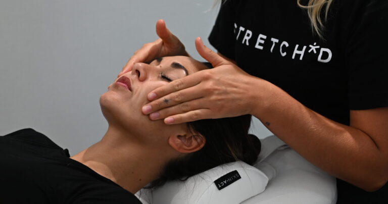 A Stretch*d team member massages a client's cheeks and temples as they lie on a cushioned headrest