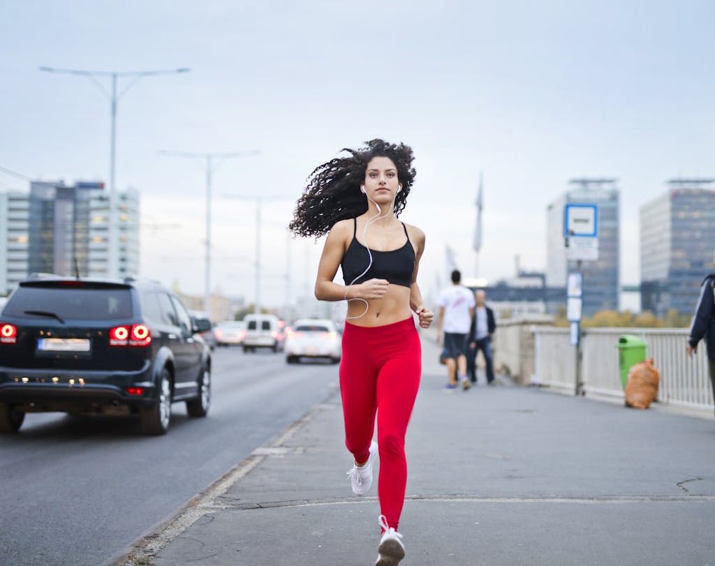 A fit woman training for a marathon with curly hair runs on a city street, enjoying music and staying healthy.
